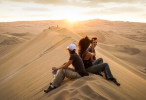 Friends enjoying magical desert sunset on top of sand dune adjacent to famous Huacachina oasis, near