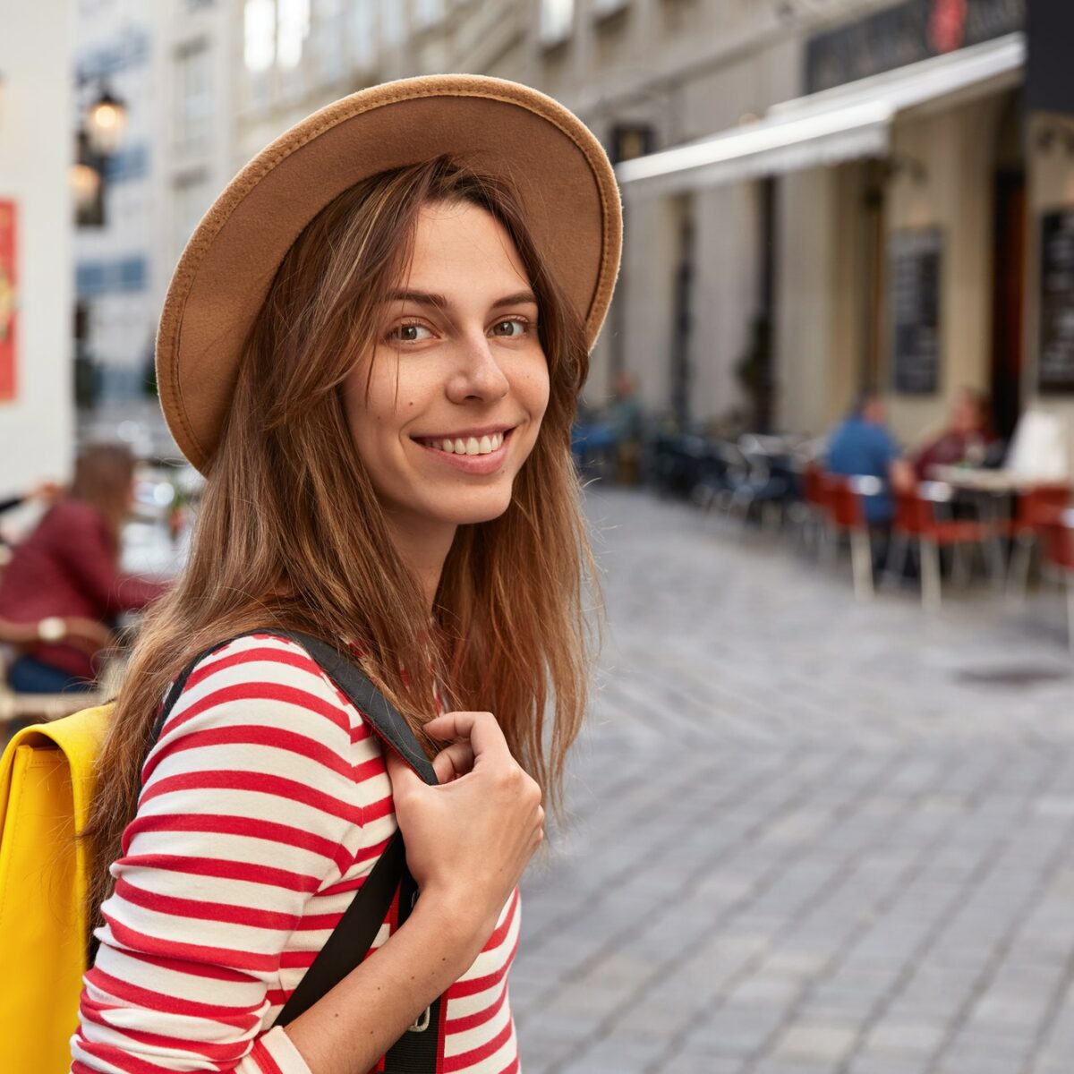 Outside shot of mirthful European tourist strolls across city streets, carries rucksack, wears brown